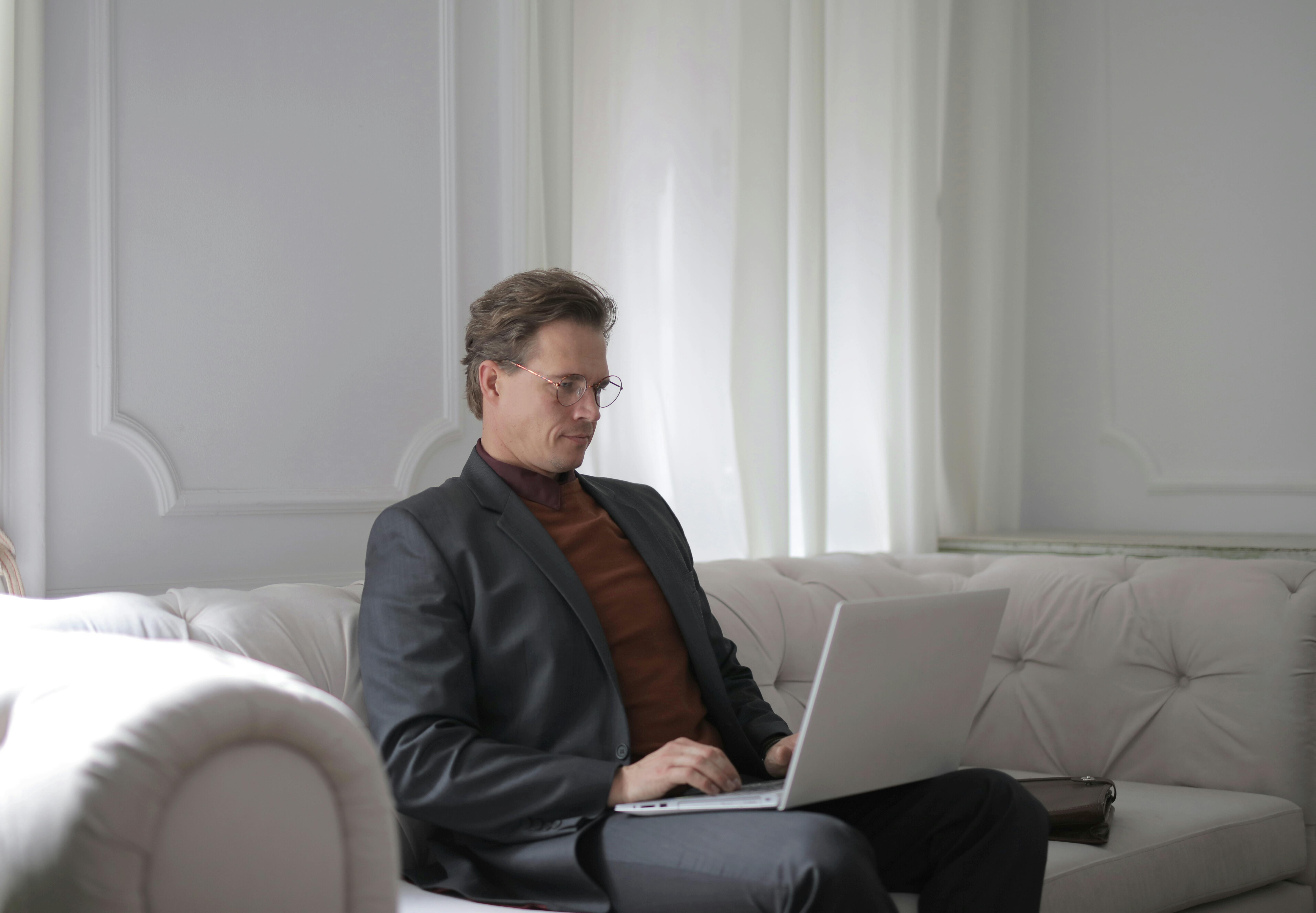 Мужчина сидит на белом диване за ноутбуком · Бесплатные стоковые фото